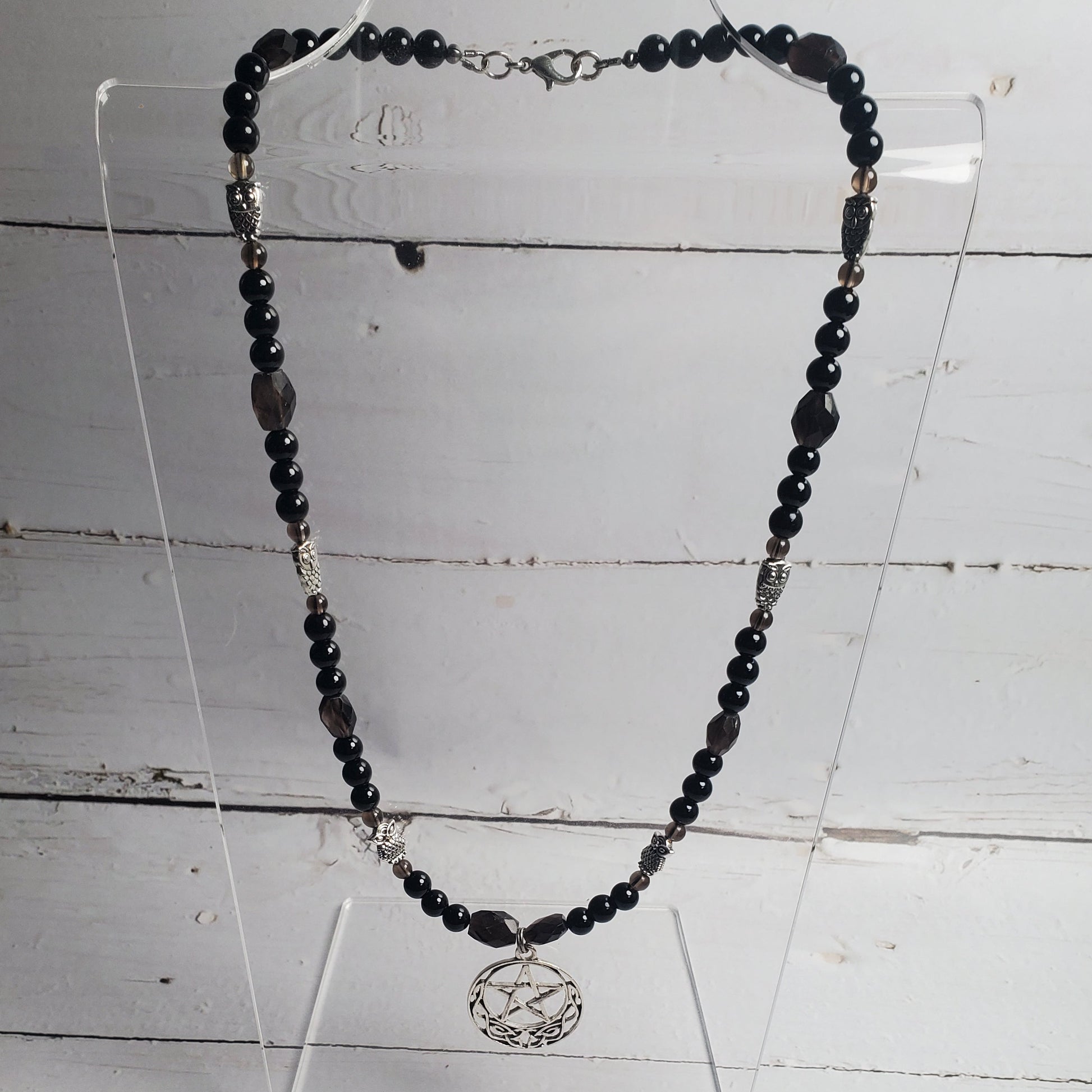 A beaded necklace with dark black and gray beads, silver spacer beads, and a silver-toned circular pendant, displayed on a clear acrylic stand against a white wood plank background.
