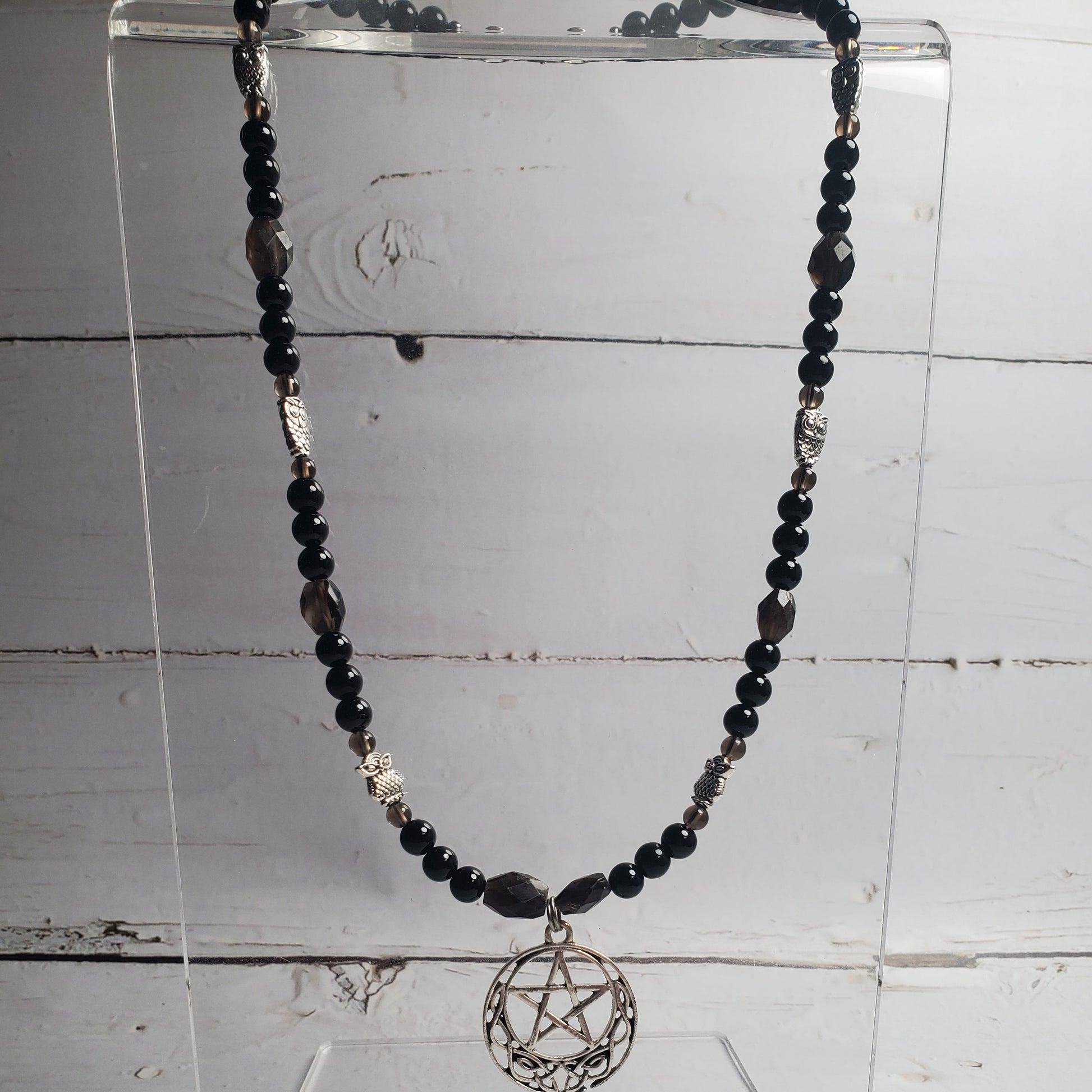 A beaded necklace with black and dark gray beads, silver-toned spacer beads, and a circular pentagram pendant, displayed on a clear acrylic stand against a white wood plank background.