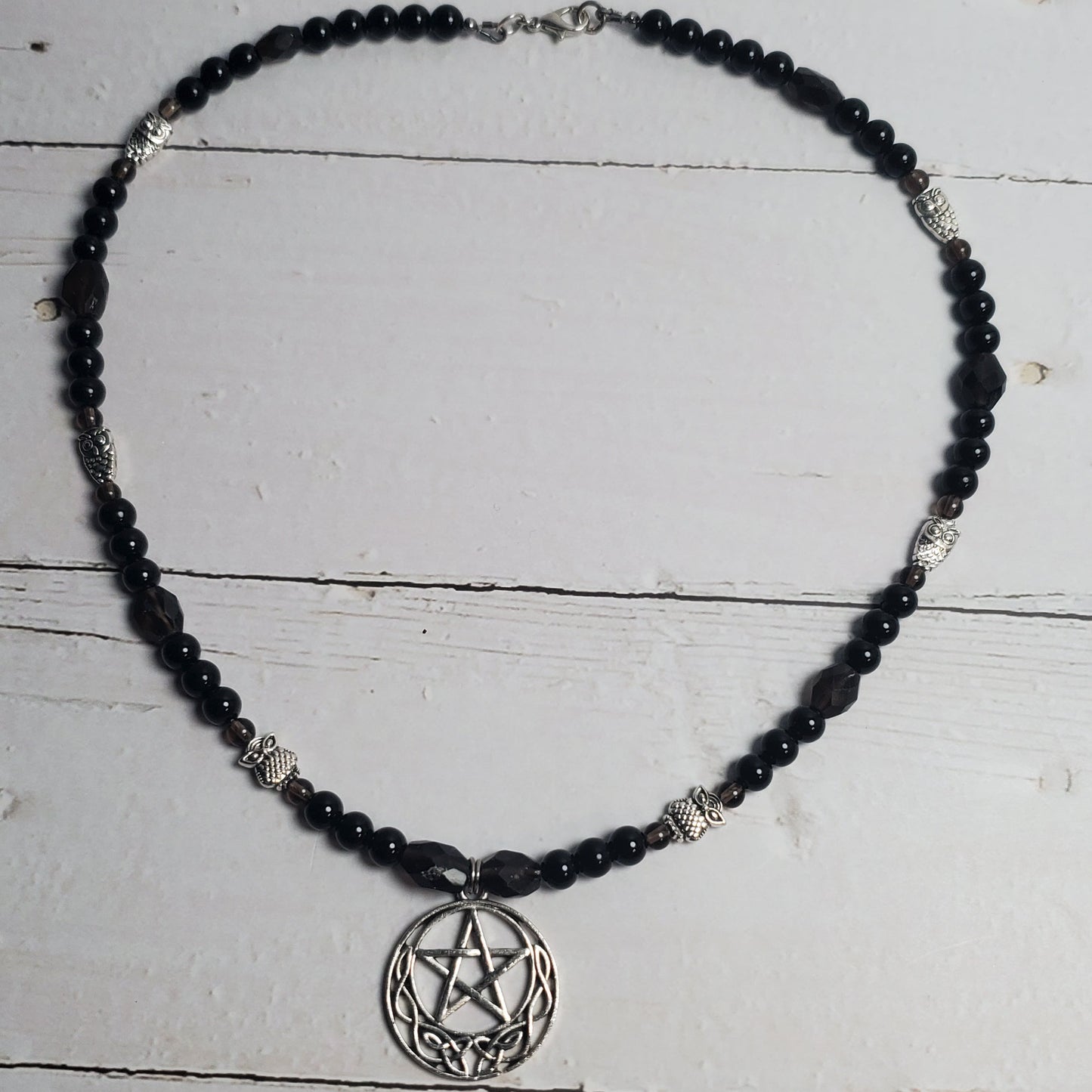 A beaded necklace featuring black faceted beads and silver-toned metal accents, including a circular pentagram pendant, displayed on a white wooden plank surface.