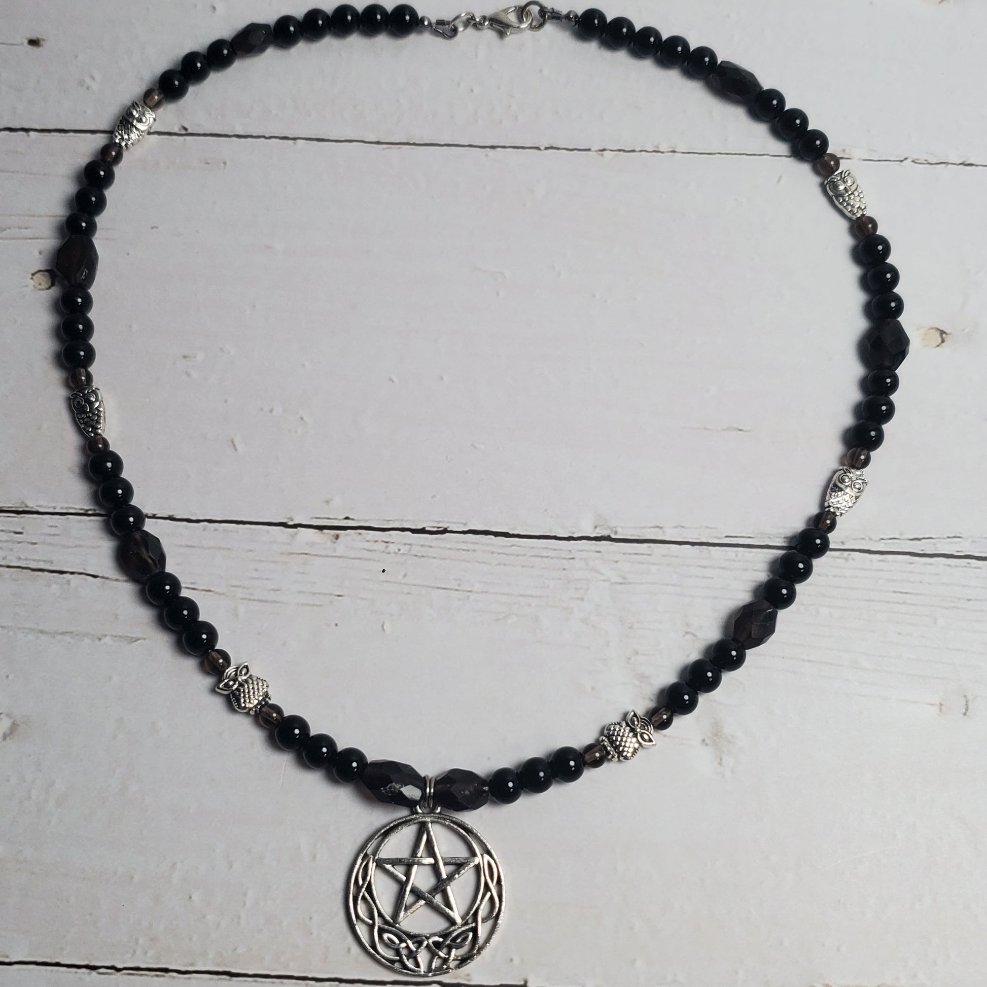 A beaded necklace featuring black faceted beads and silver-toned metal accents, including a circular pentagram pendant, displayed on a white wooden plank surface.