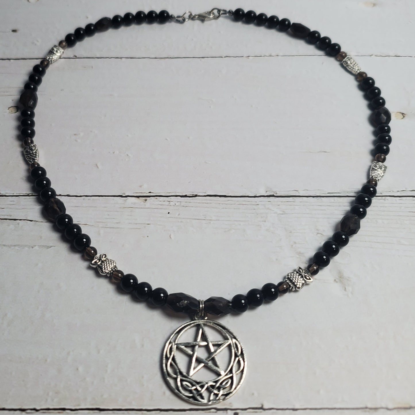 A beaded necklace featuring black faceted beads, silver-toned spacer beads, and a circular silver pentagram pendant, displayed on a white wooden plank surface.