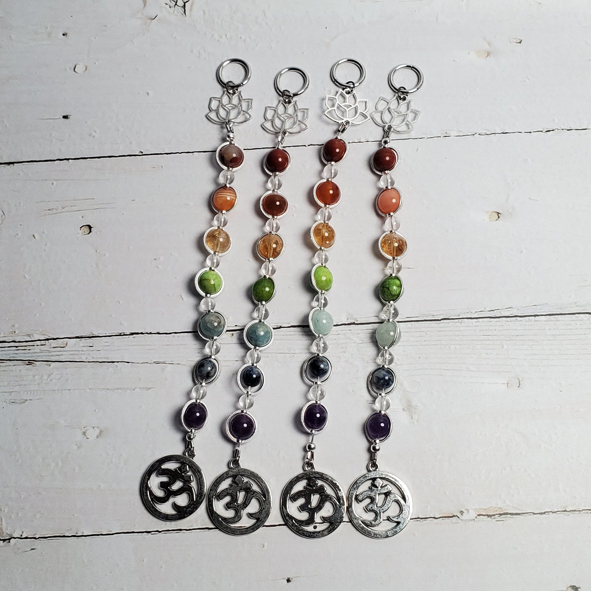 Four beaded chakra wall hangers with silver Om symbols, displayed on a white wooden surface.