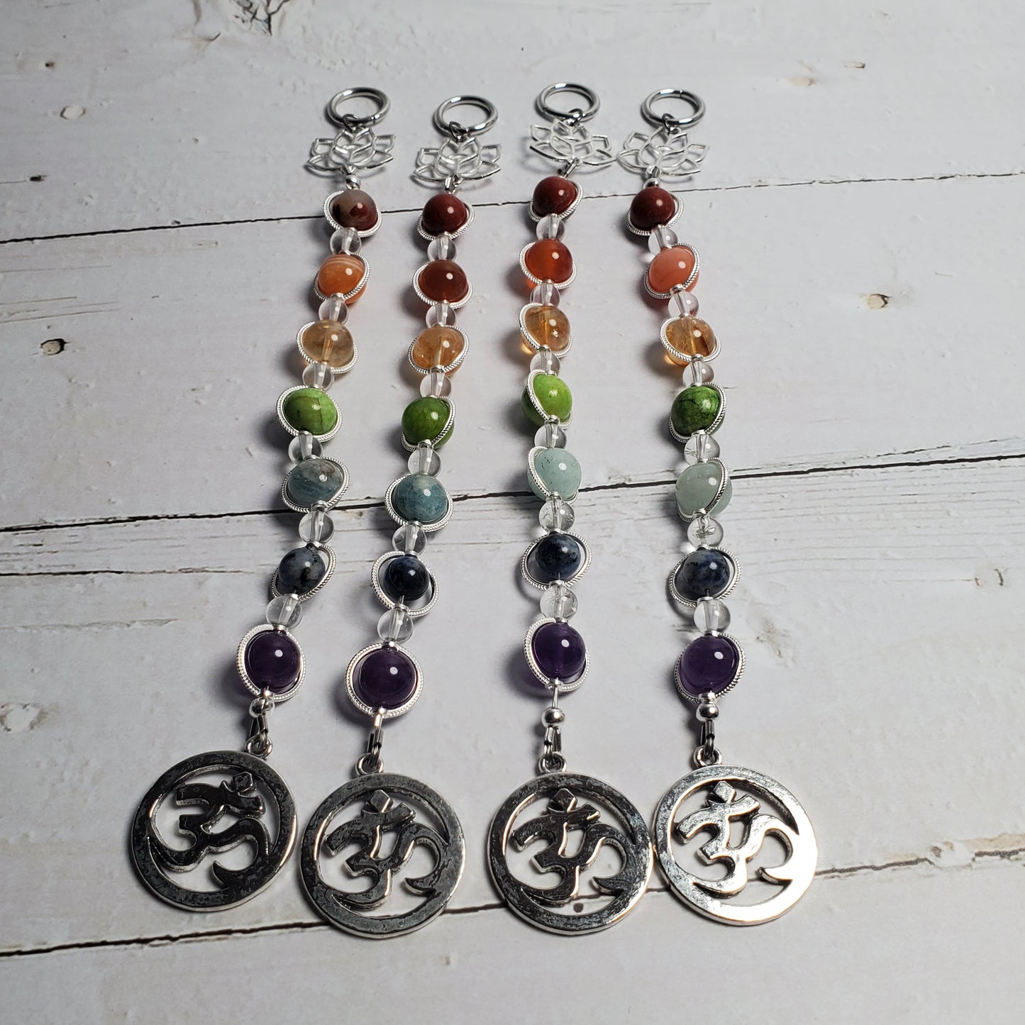 Four beaded spiritual stitch wall hangers with silver Om symbols, arranged vertically on a white wooden surface. Each tag features a string of multicolored gemstone beads (red, orange, green, blue, purple) attached to a silver clasp and a circular silver Om charm at the bottom.