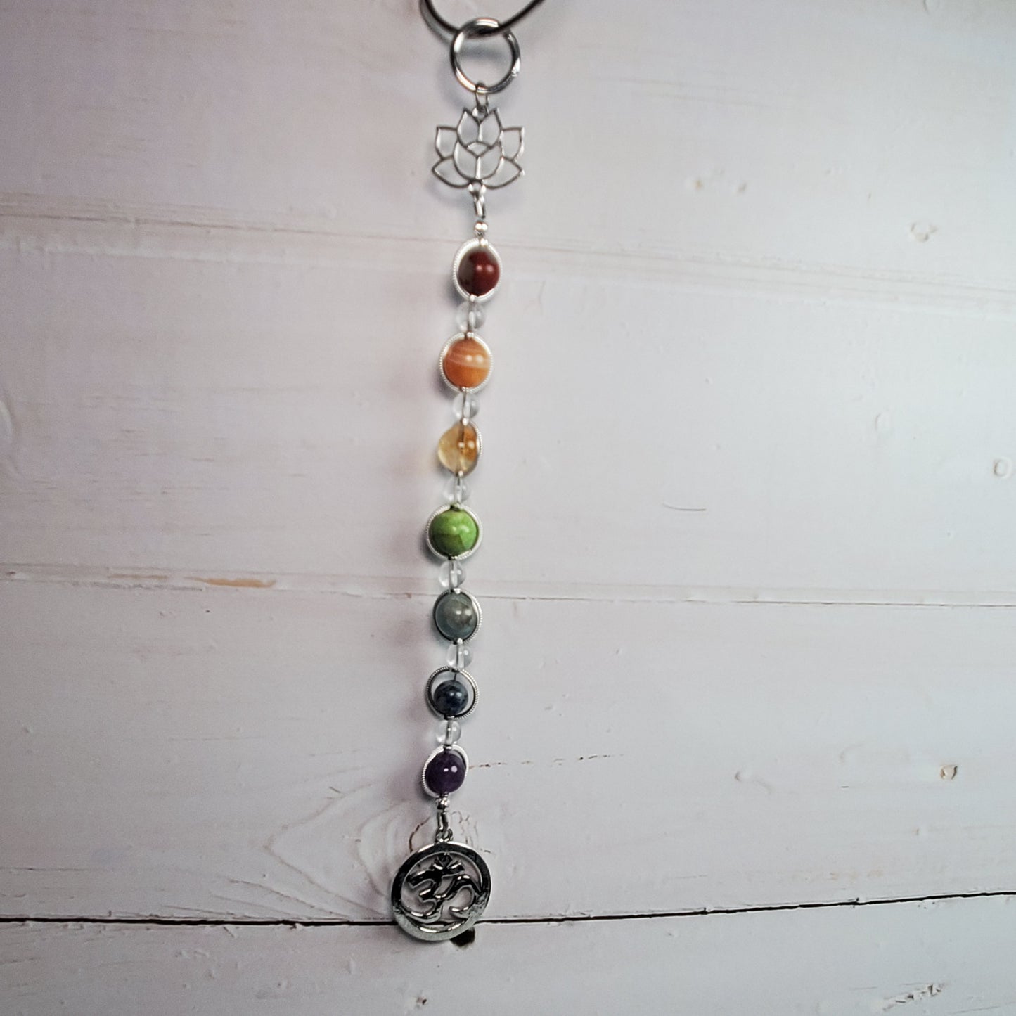 A beaded chakra wall hanger with silver-toned metal charms, including a lotus flower, hanging against a white wooden plank background.