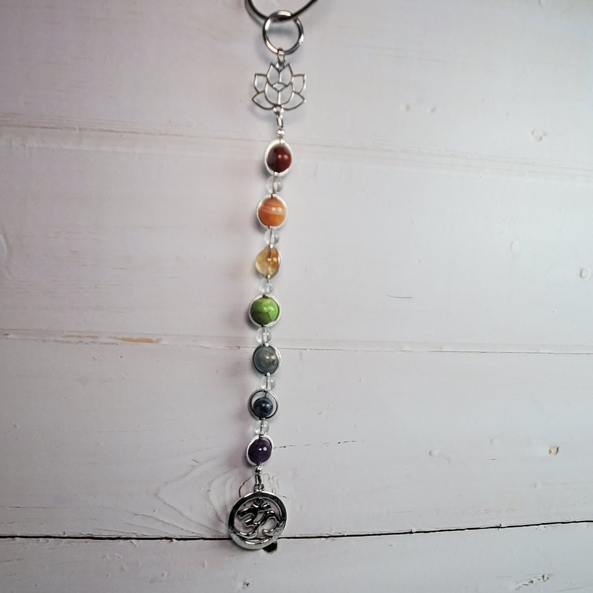 A beaded chakra wall hanger with silver-toned metal charms, including a lotus flower, hanging against a white wooden plank background.
