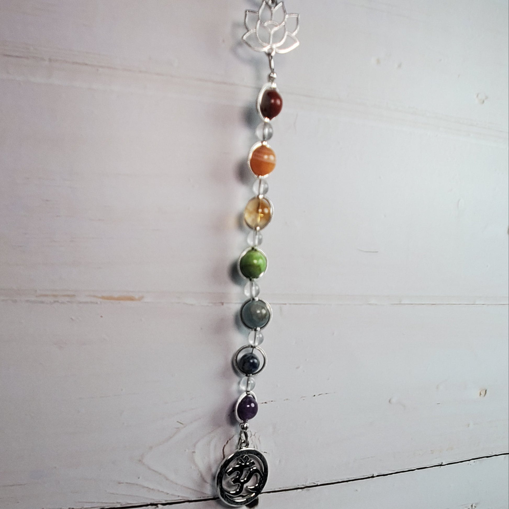 A vertical chakra bead hanging ornament with silver-toned metal links, colorful gemstone beads, a lotus flower at the top, and a circular Om symbol charm at the bottom, displayed against a white wooden wall.