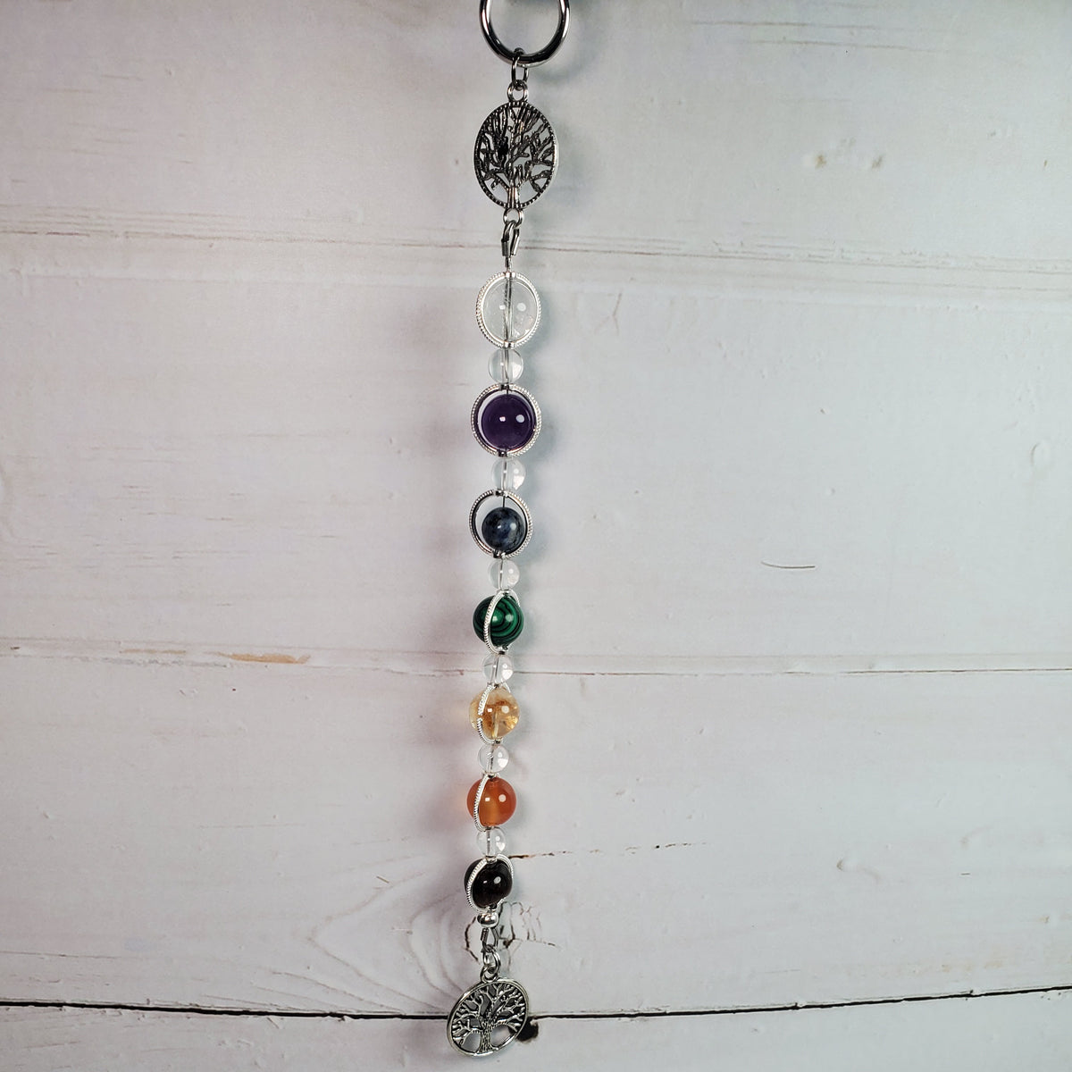 An intention hanging ornament with silver-toned metal charms and clear beads, featuring colored gemstone beads in purple, blue, green, yellow, orange, black, and red, hanging against a white wooden plank background.