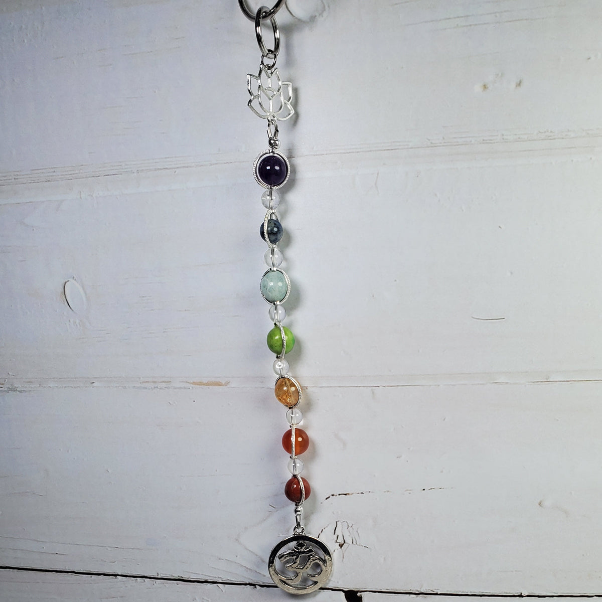 A beaded chakra wall hanger with a silver-toned top, clear crystal, purple, green, orange, and red beads, and a silver coin pendant, hanging against a white wooden plank background.