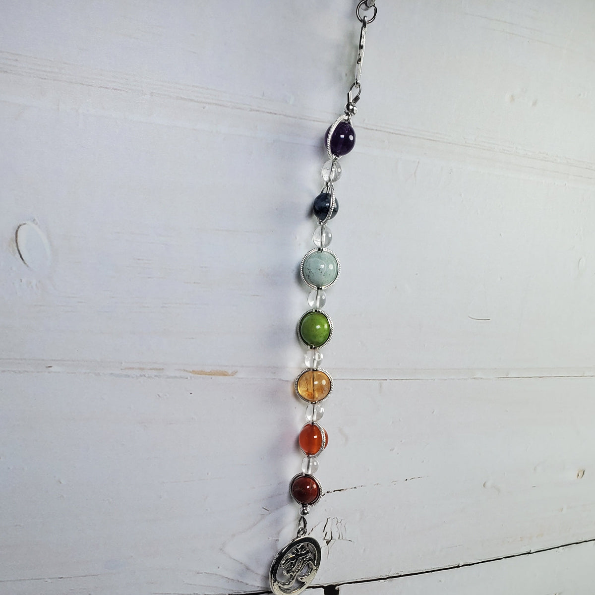 A beaded chakra wall hanger with seven colorful gemstone beads and a silver-toned metal pendant, hanging against a white wooden plank background.