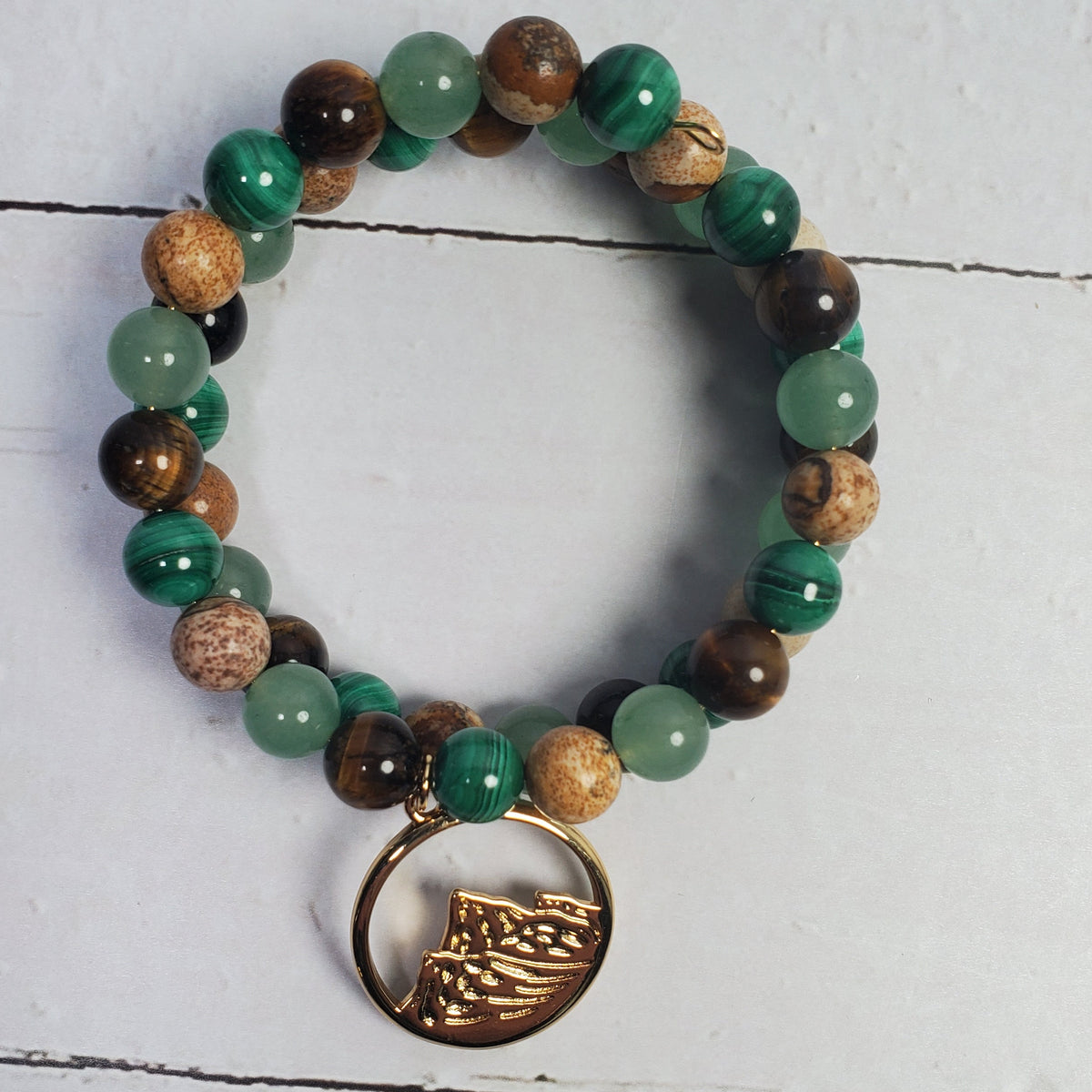 A beaded memory wire bracelet featuring green and brown stones, including tiger's eye, malachite, and wood beads, with a bronze-toned circular mountain charm, displayed on a white wood background.