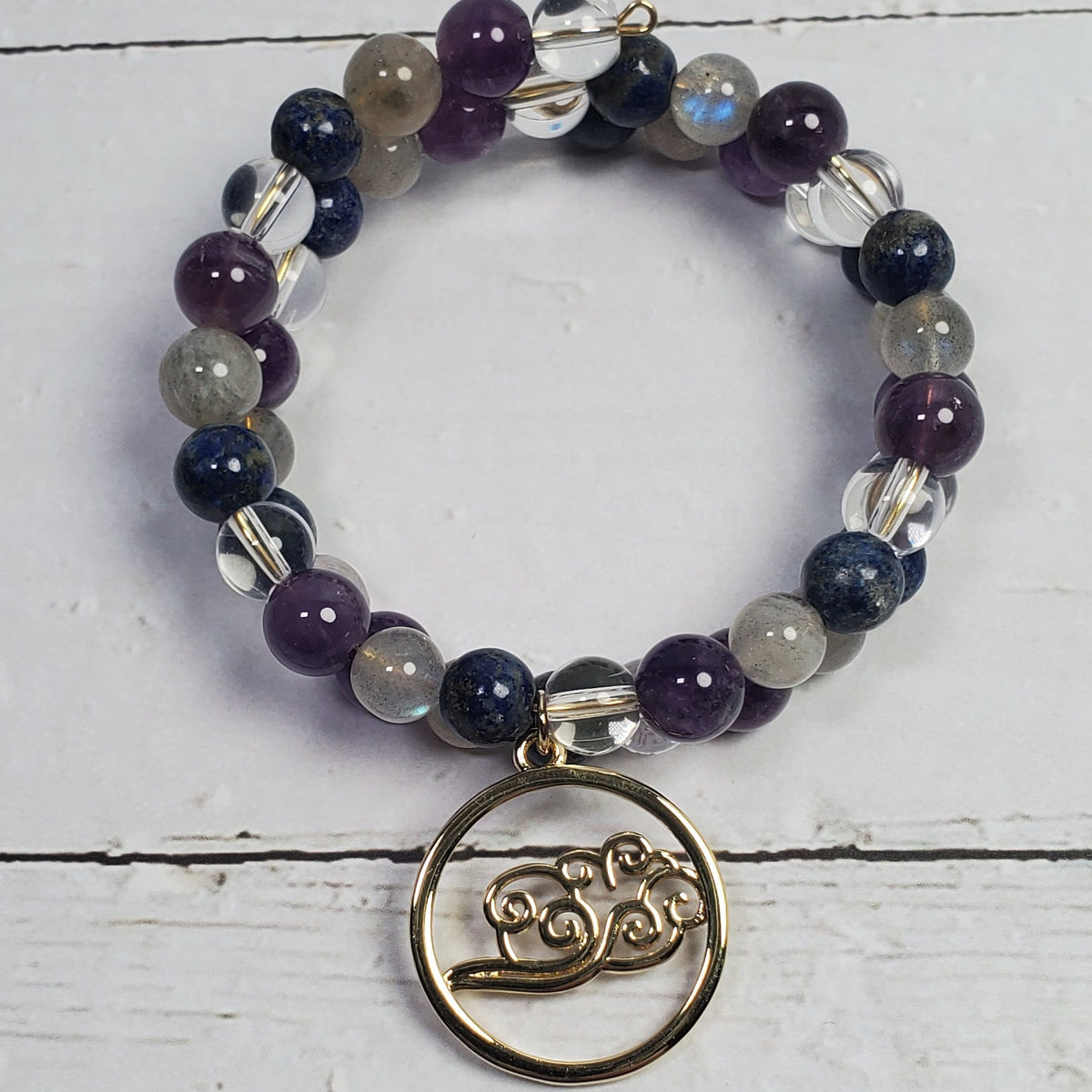 A beaded memory wire bracelet featuring purple, navy, gray, and clear beads, with a bronze-toned Om symbol charm hanging from a circular ring, displayed on a white wooden background.