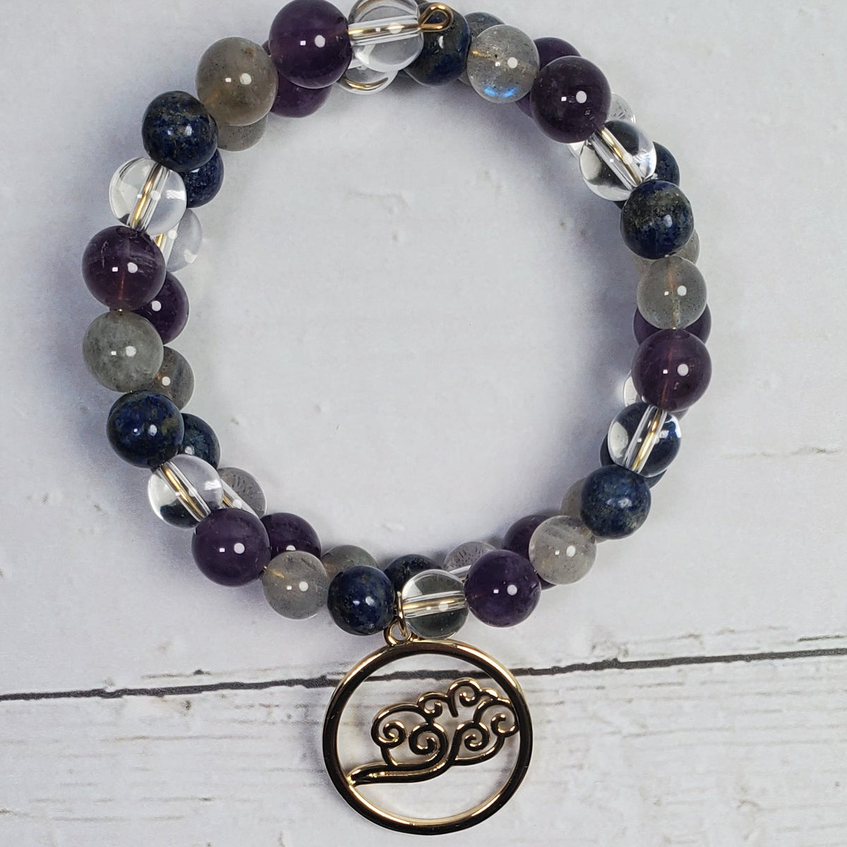 A beaded memory wire bracelet with purple, gray, and clear faceted beads, featuring a small circular metal charm engraved with a bicycle, displayed on a white wooden surface.
