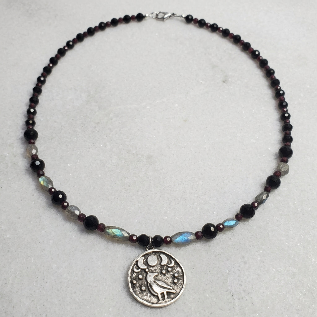 A beaded necklace with dark faceted beads, gray stone beads, and a silver-toned round pendant featuring a bird and moon design, displayed on a white wood plank background.