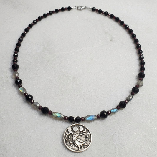 A beaded necklace with dark faceted beads, gray stone beads, and a silver-toned round pendant featuring a bird and moon design, displayed on a white wood plank background.