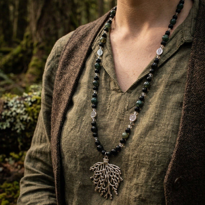 A beaded necklace with dark green and black round beads and a silver-toned tree-of-life pendant, displayed on a clear acrylic stand against a white wood plank background.