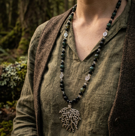 A beaded necklace with dark green and black round beads and a silver-toned tree-of-life pendant, displayed on a clear acrylic stand against a white wood plank background.